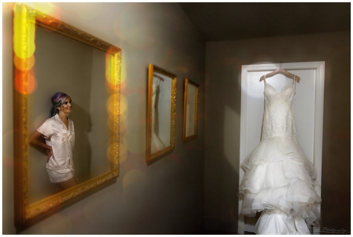 wedding dress hangs on door as bride looks on in reflection image