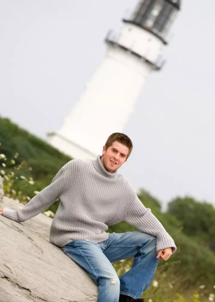 boy in gray sweater with lighthouse at senior pictures at the beach in maine