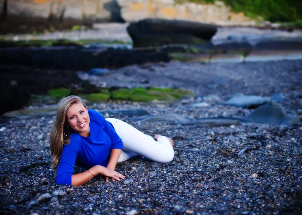 girl in blue shirt on stones for senior pictures at the beach in maine