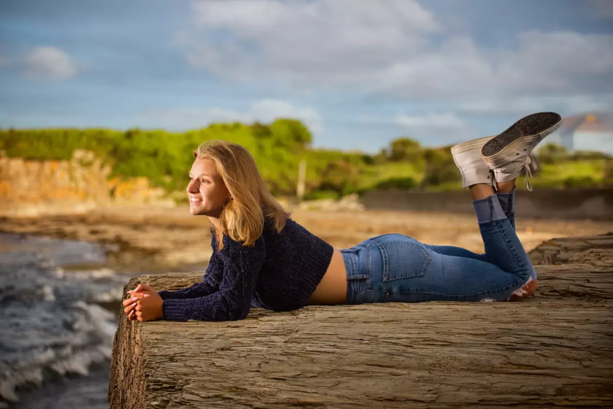 girl in jeans and sweater lying on rock over ocean during professional senior photos in maine at the beach
