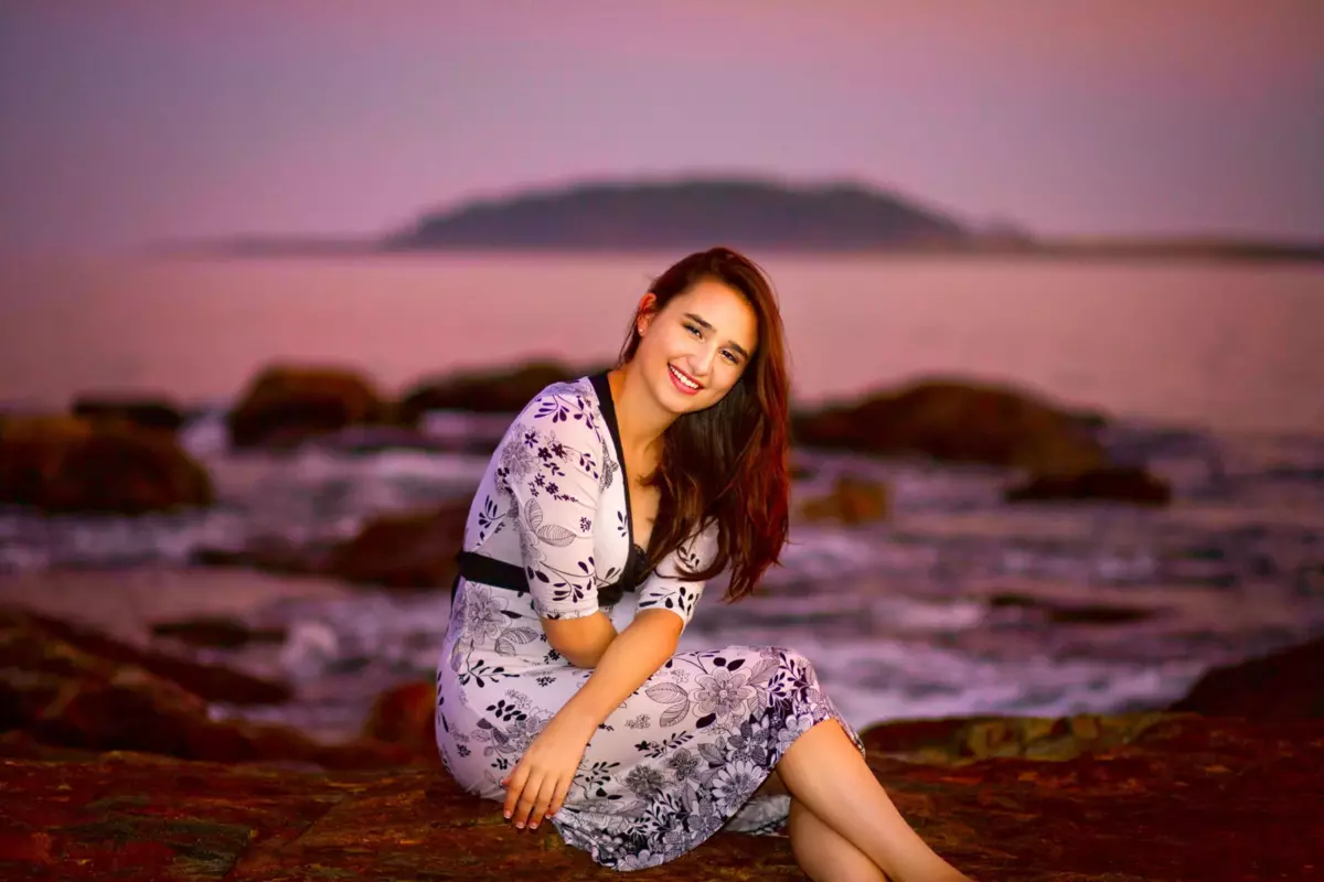 high school girl in dress seated on driftwood at professional senior photos in maine at the beach