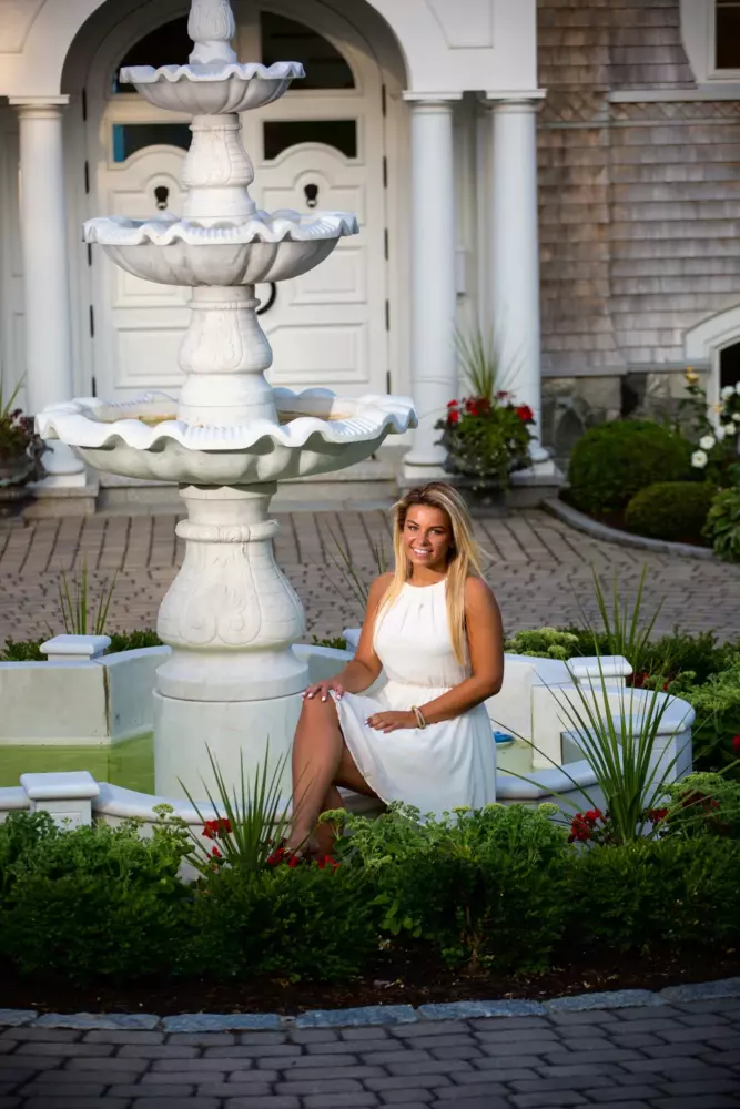 high school girl in white dress sits at fountain at entry to home in south portland, maine senior pictures