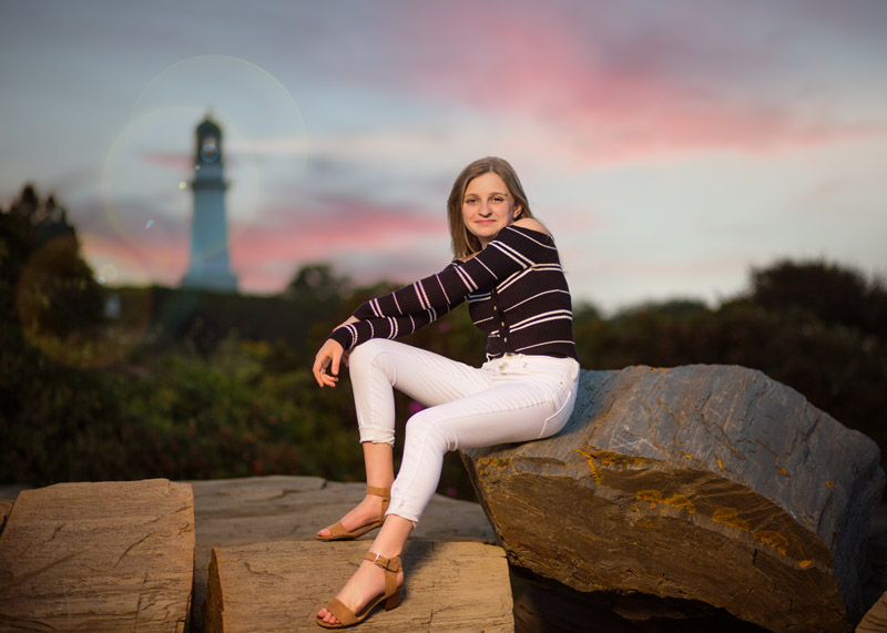 Senior portrait taken in Maine at sunset with lighthouse in distance for high school yearbook pictures.